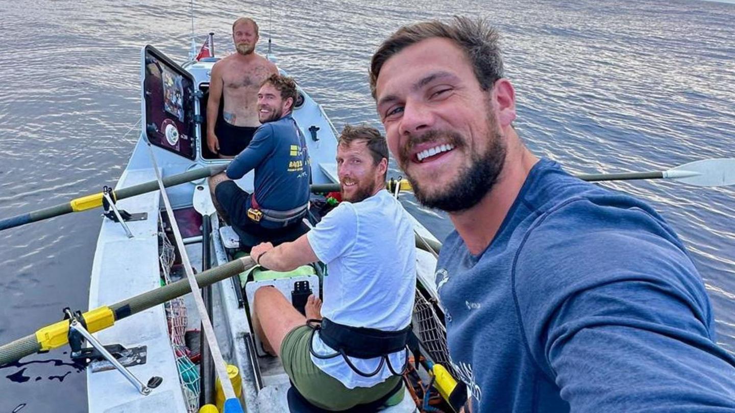 The four crew members of United We Conquer smiling while rowing their boat in the open ocean. One of them is holding the camera for a selfie. One is standing up.