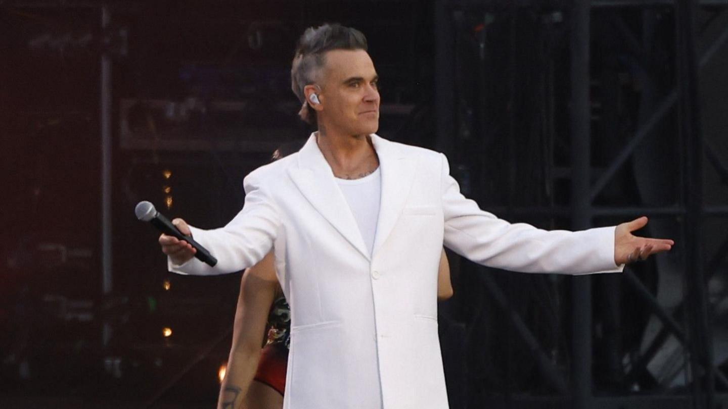 Robbie Williams is dressed in an all-white suit on a stage in front of thousands. There are backing dancers dressed in sparkly costumes behind him.