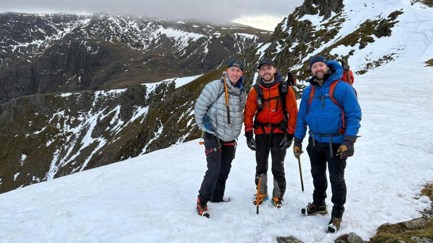Mae tri dyn mewn cotiau yn sefyll ar ben crib greigiog wedi'i orchuddio ag eira gyda chopa mynydd y tu ôl iddyn nhw