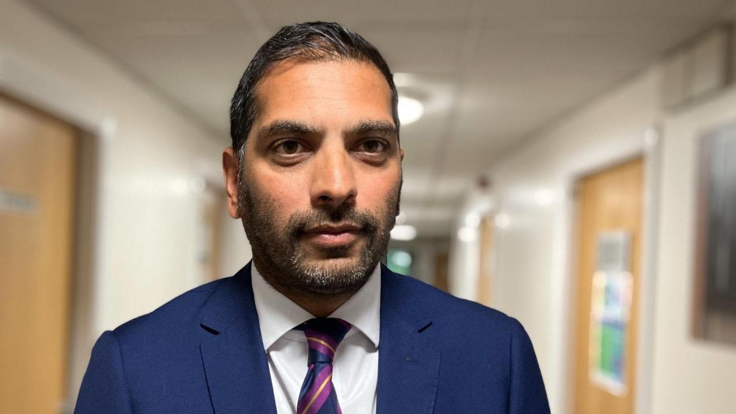 Steve Russell is in a navy suit standing in a hospital. He is wearing a striped purple, yellow and navy tie.