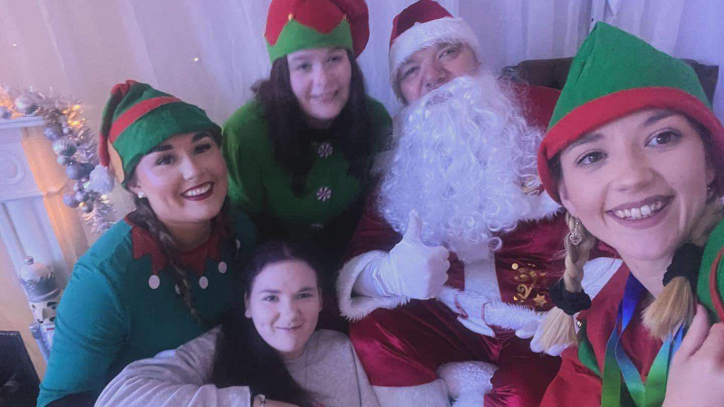 A group of women dressed as Christmas elves pose with a man dressed as Father Christmas. Santa has a bush white beard, red jacket with white fur trim and a red and white hat. He is doing a thumbs up to the camera with a white gloved hand. The elves are wearing green tops and green and red pointed hats.