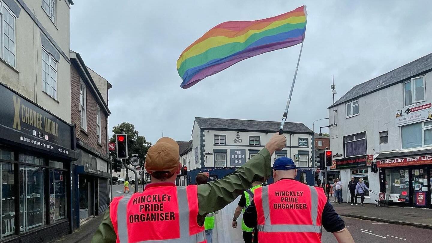 A rainbow flag waves in the air, with two individuals in the foreground wearing pink hi vis jackets with "Hinckley Pride Organiser" written on them.