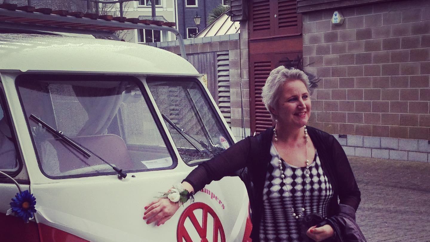 Becky Rowland stands in front of a vintage VW camper can in red and cream - she wears a monochrome dress and a fascinator on her head