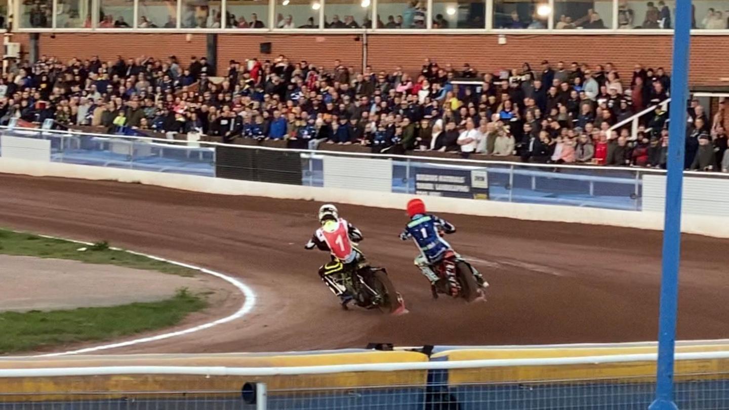 Two speedway riders drifting on the bend while spectators watch from stands