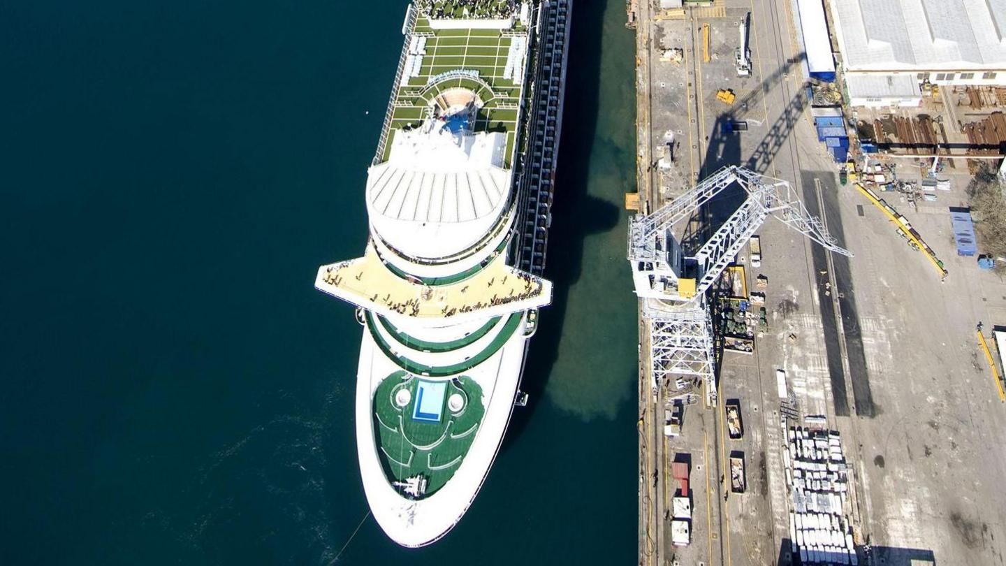 Ventura looking down onto the front of the ship with Southampton's dockside to the right with vehicles and a crane on the edge. Looking down on the ship you can see swimming pools and a green area. There is a small crowd of people standing looking out to sea.