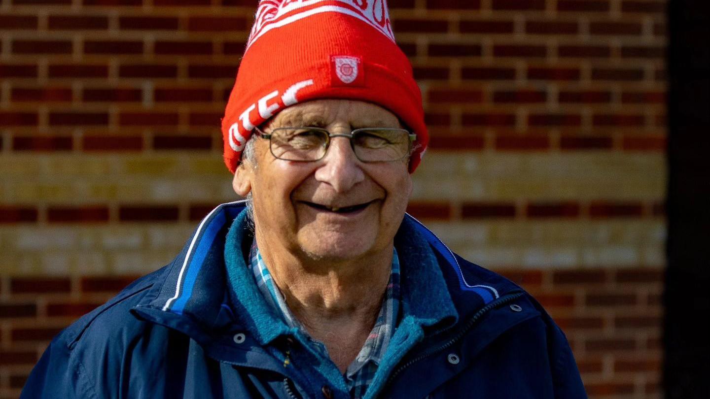 A man wearing a blue anorak, a red hat and a pair of glasses, smiling at the camera in front of a brick wall