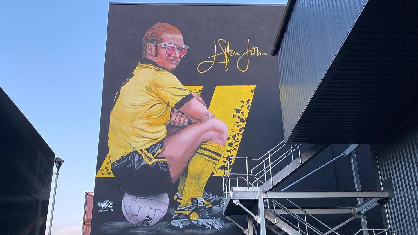 A mural of Sir Elton John on the side of a football stadium. He wears a yellow football shirt and black shorts while sitting on a football. He has pink glasses and short ginger hair.