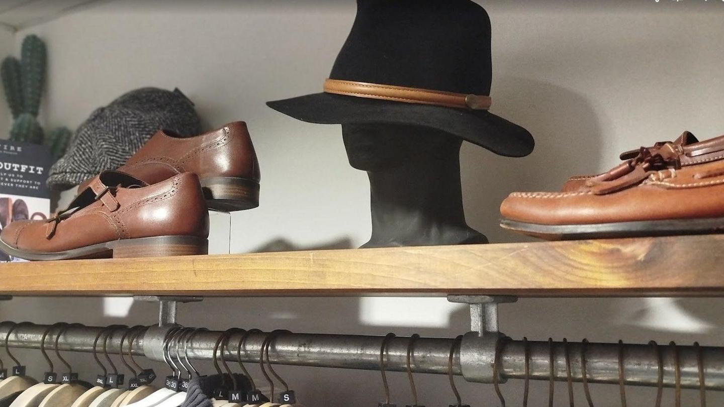 Brown shoes and a black fedora sit on a shelf above a clothes railing. 