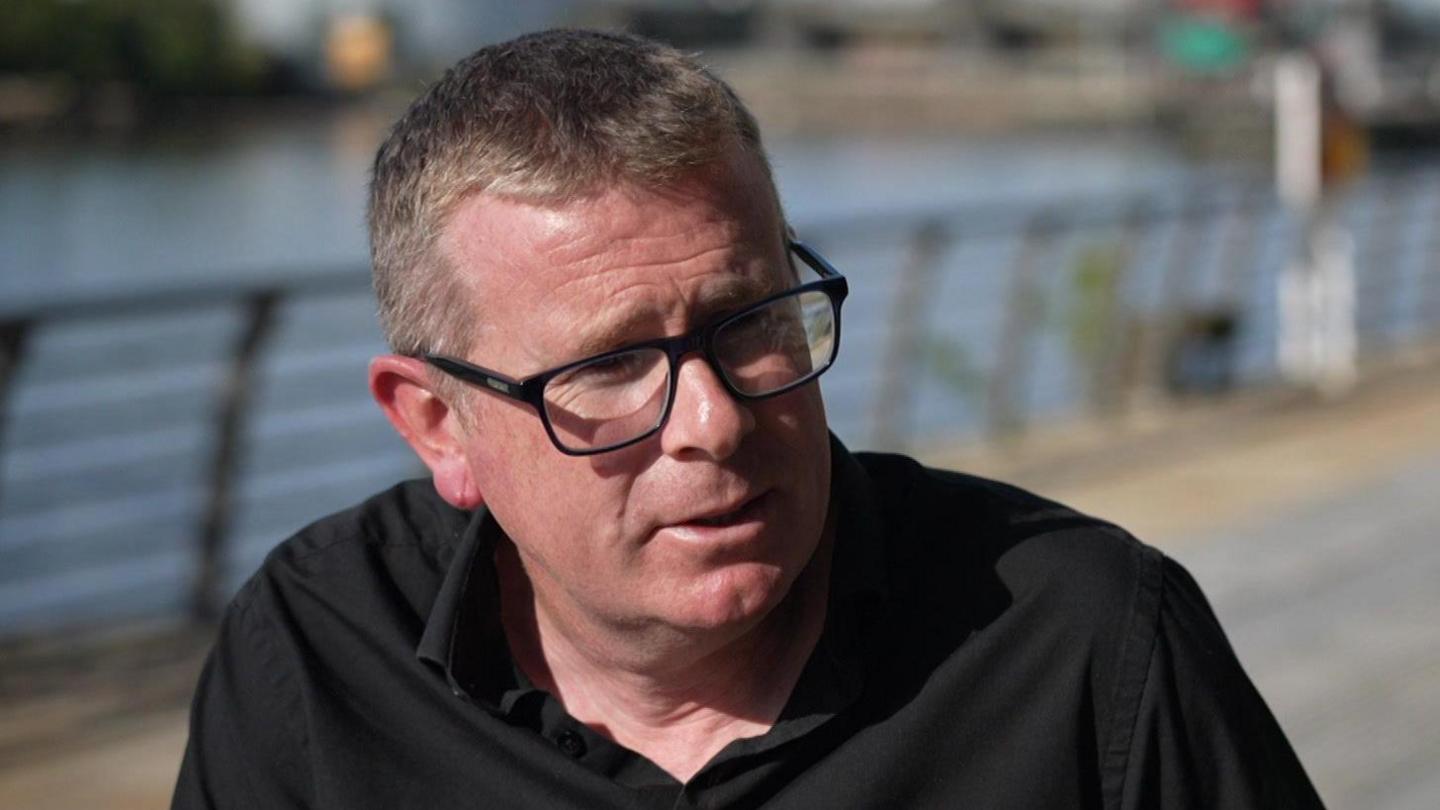 Dr John Mulligan wearing glasses and a black shirt sitting on a bench with railings and water in the background