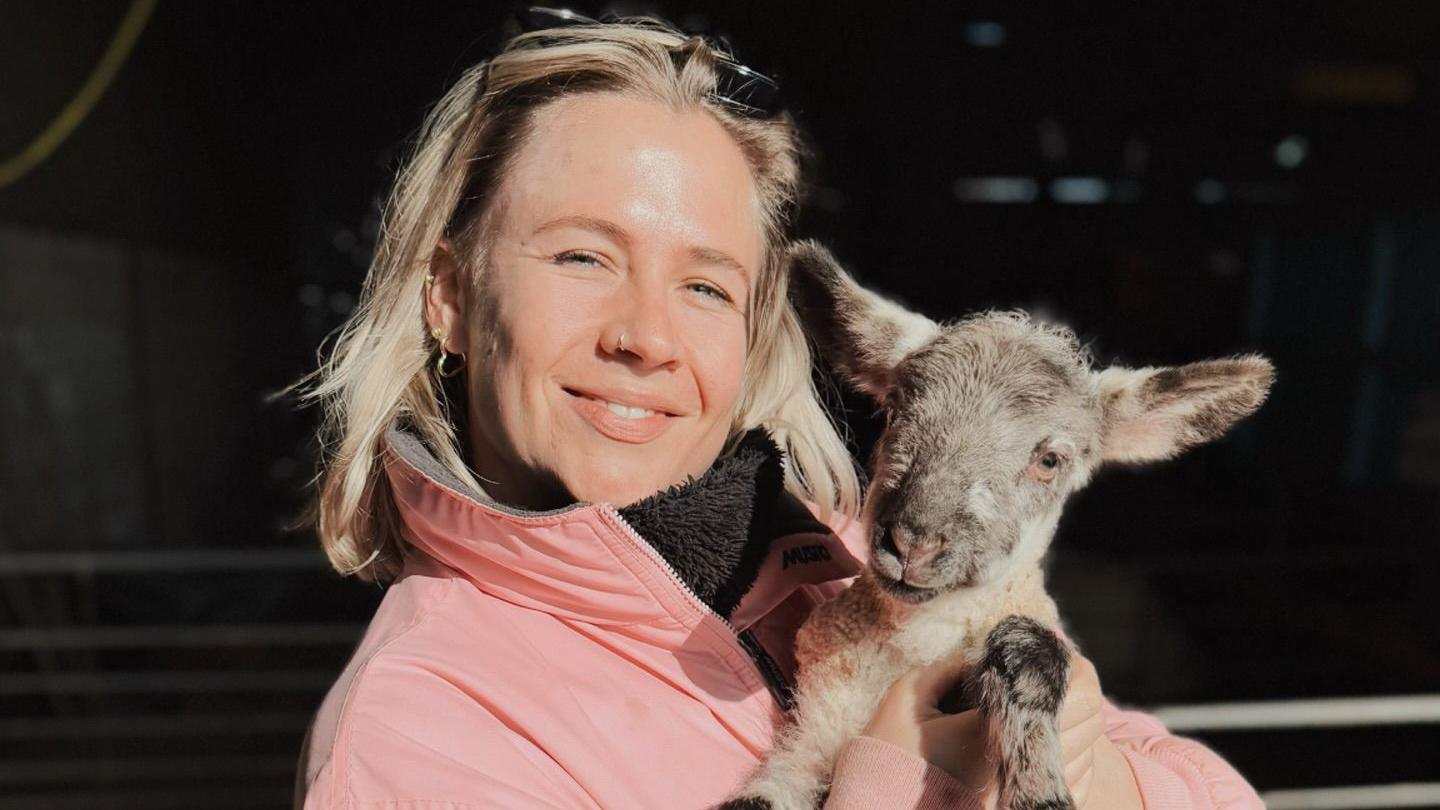 A woman looking at the camera in a pink coat holding a lamb