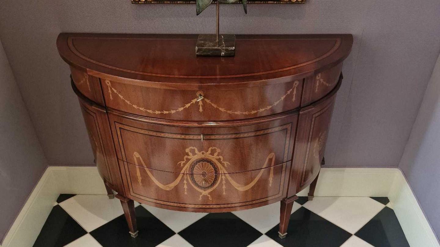 A demilune commode with a top inlaid with satinwood and ebonised strung banding, below are three drawers with conforming inlay together with bell flower swags, drapery and an oval fan medallion, on tapered square legs ending in brass capping.