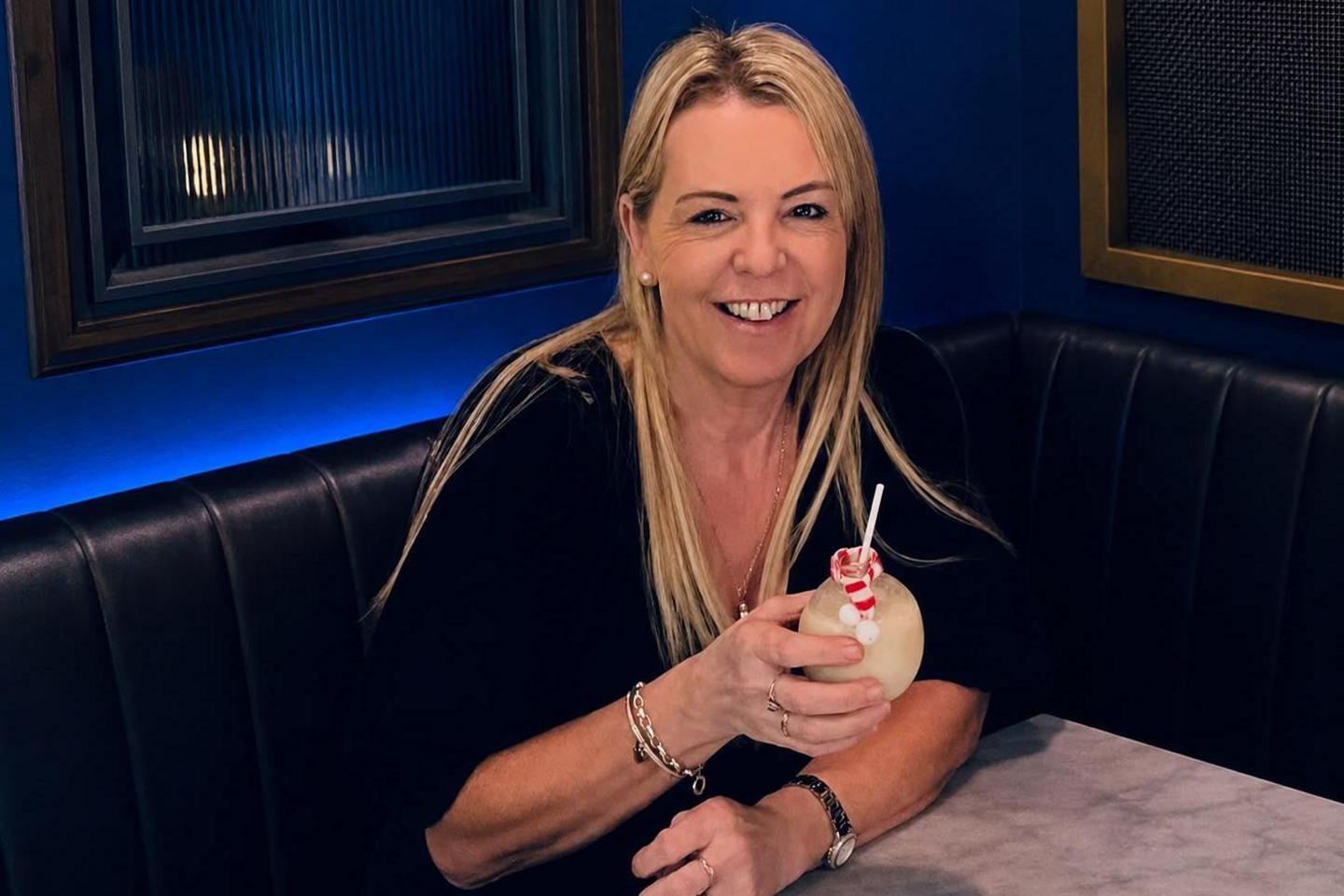 A smiling Deb Colley has long blonde hair and is wearing a black dress and holding a cocktail