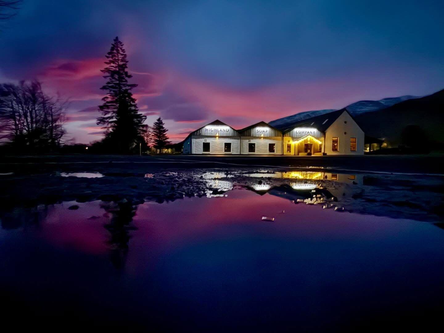 A blue and pink coloured sunrise over a loch with four white buildings on the waters edge