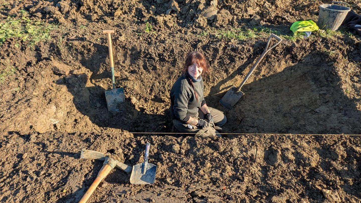 Woman sat inside a ditch dug into earth at Historic England's excavation site, near Willersey. She is wearing black jumper and trousers and has red hair.