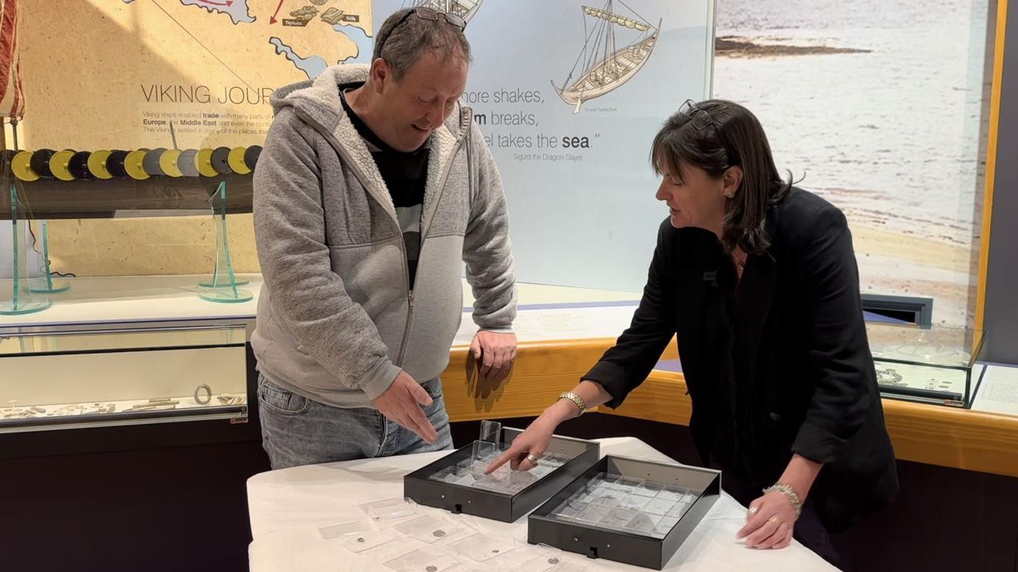 David O'Hare and Allison Fox looking at the silver coins, which are in plastic cases in two black drawers on a table in the Viking Gallery in the Manx Museum. They are standing in front of panels displaying information about the Viking age and glass display cases holding other Viking treasure.