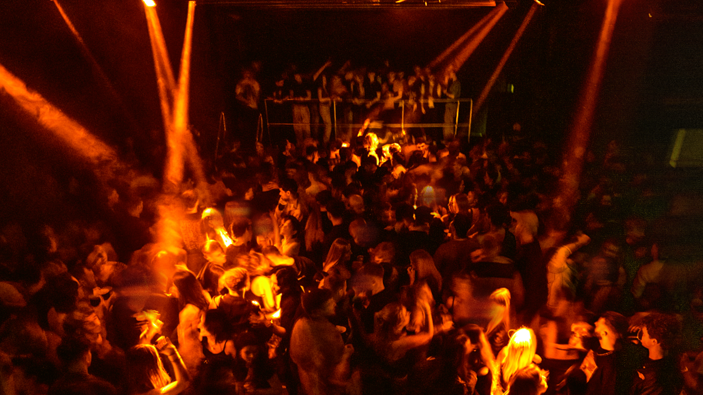 A busy night club dancefloor. There are orange lights shining down on a crowd of people dancing.