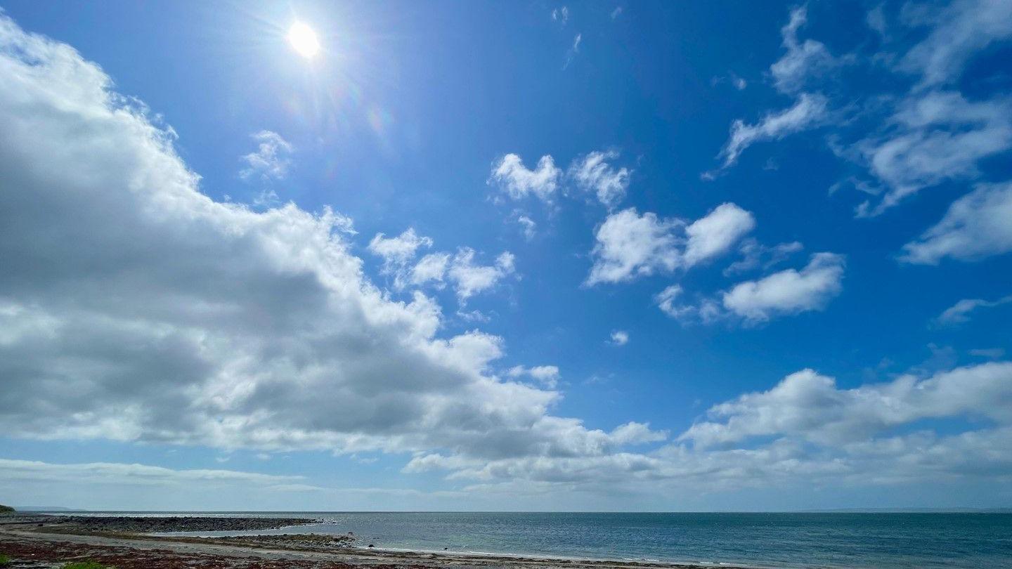 The sun shines in a blue sky, with patchy cumulus clouds, above the sea