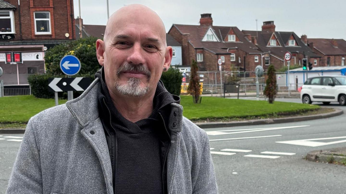 Alan Jones is pictured in front of a traffic island. There are shops, houses and cars behind him. He is a middle-aged man with a bald head and a grey goatee beard. He is wearing a dark grey hooded top over a light grey coat. He is smiling at the camera.