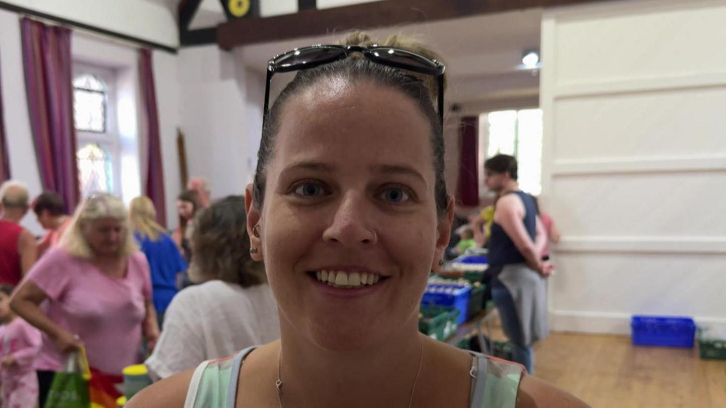 Victoria Henstridge smiles for the camera, with a bustling community larder behind her in a large village hall. She is wearing a grey top and had sunglasses on her head.