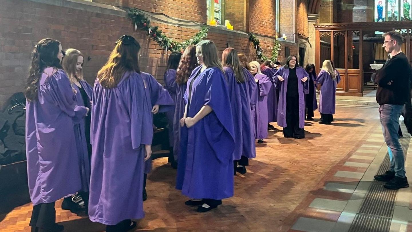 The group of young women singers in a church wearing purple robes. The choir's founder, Duncan Rutherford, looks on from the side wearing a black jumper and blue jeans.