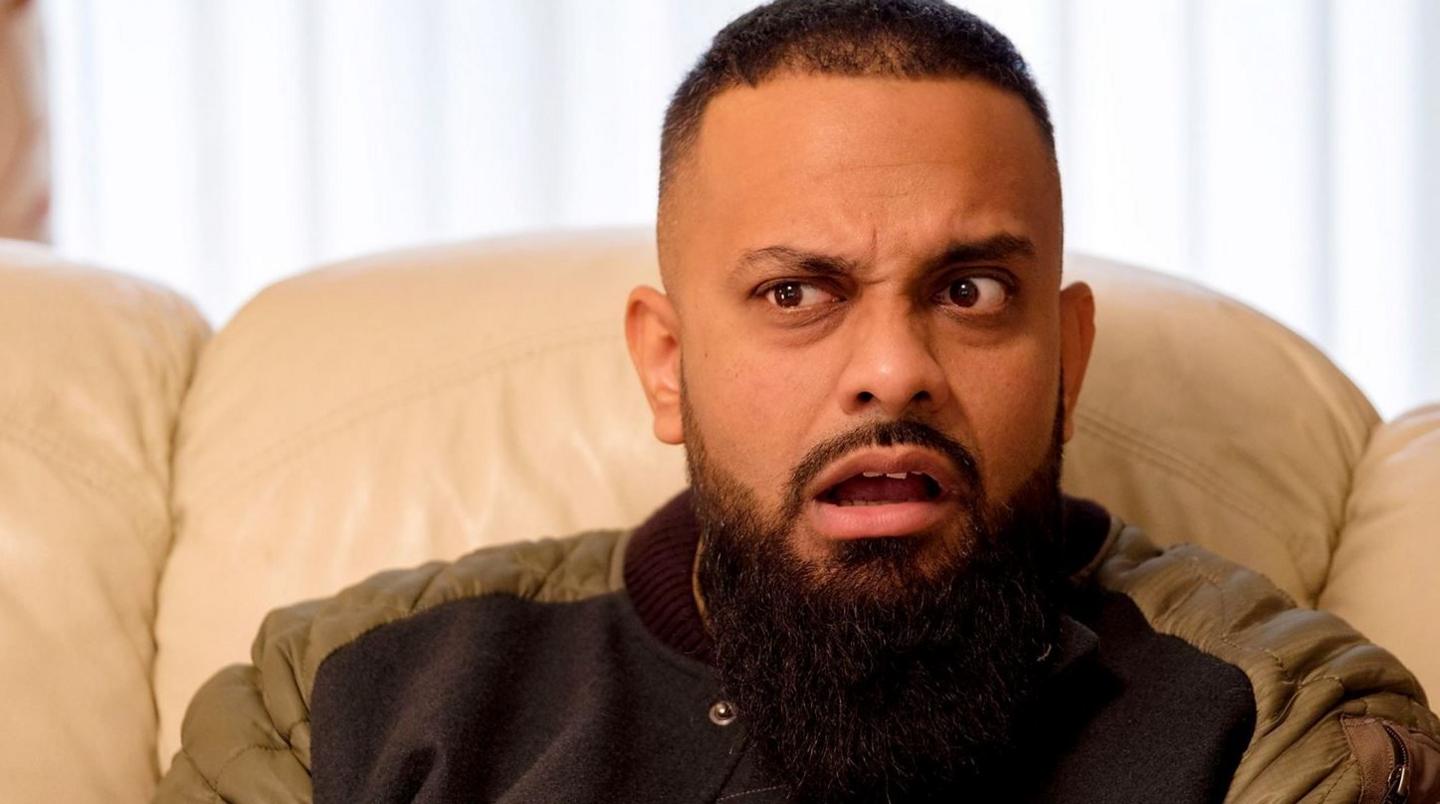 A man with short dark hair and a beard sat on a beige sofa.