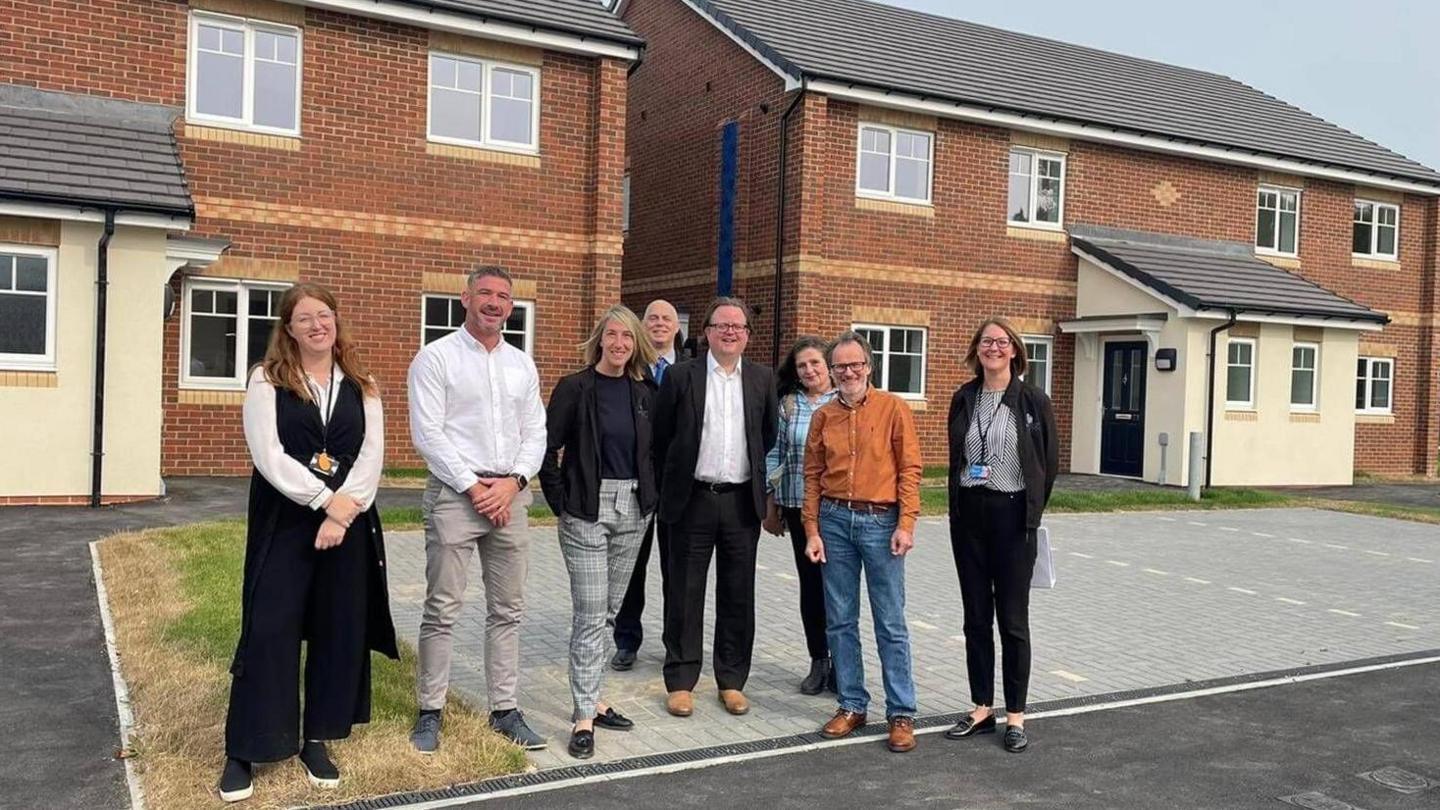 Four men and four women stand on a parking area outside two sets of newly-built semi detached properties