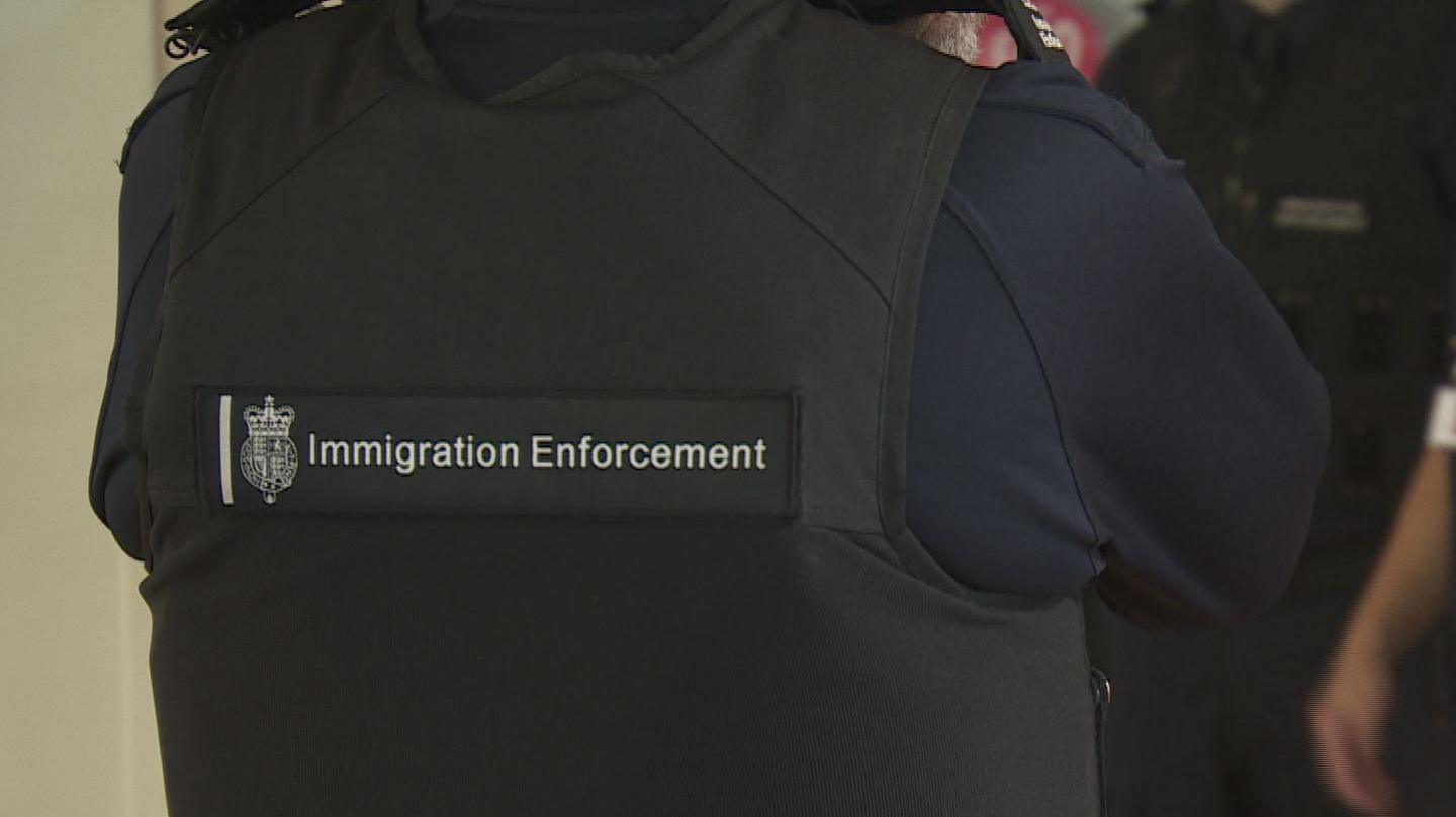 An Immigration Enforcement officer standing with their back to the camera. They are wearing a black vest with a 'Immigration Enforcement' patch on the back.