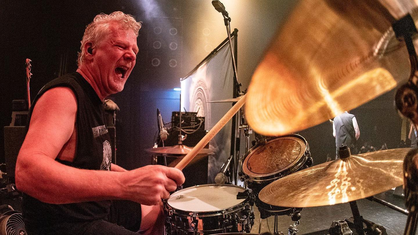 Simon Dawson playing the drums. He is wearing a sleeveless top and holding drum sticks. He is singing along to the music.