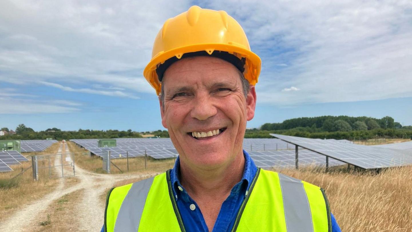 Ollie Pendered smiling at the camera in a yellow hard hat and high vis vest. Standing in front of rows of solar panels.