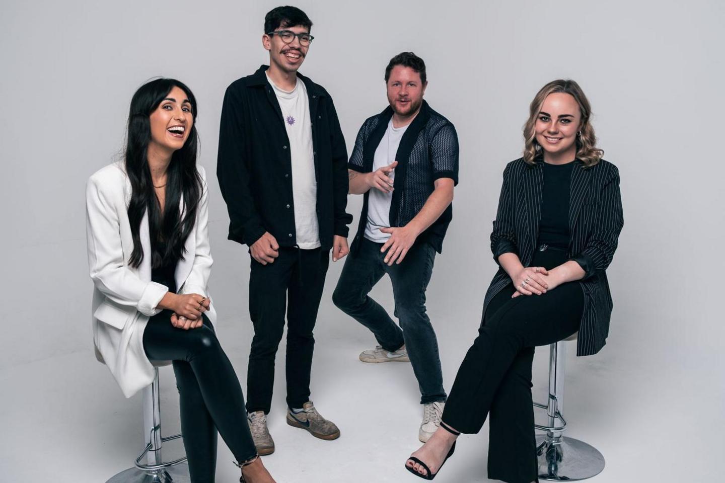 The band are striking poses in a photo studio. They are laughing and smiling. Two male members of the group are standing and the female band members are sitting on stools.