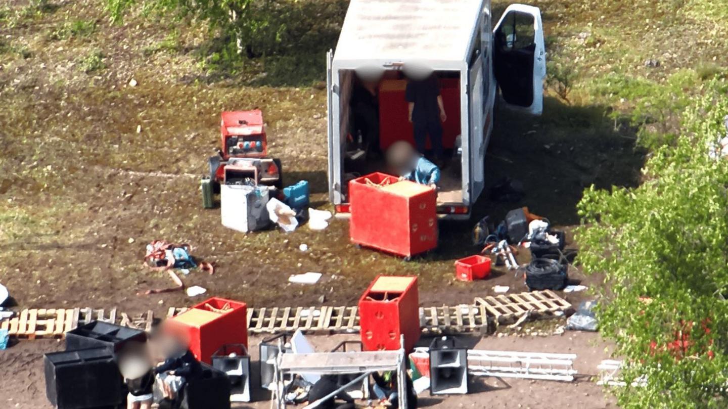 Two people standing in the back of a white van, unloading red plastic boxes full of music and staging equipment. On the ground there are various structures and debris strewn around from the event. The ground is dirty and dry and the van is surrounded by bushes.