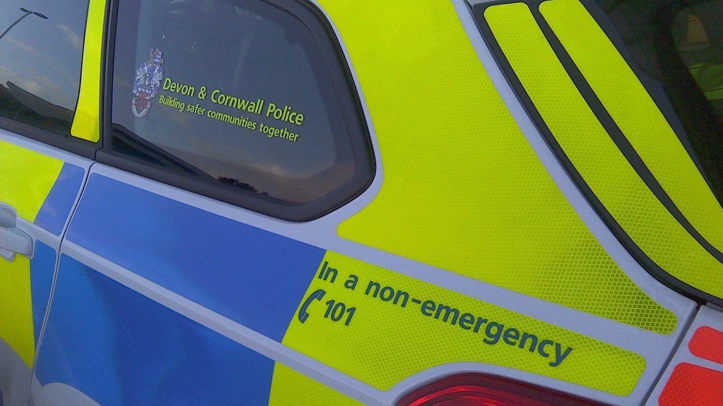 The side of a police car which has blue and yellow stickers on the side of the car. The back window says 'Devon & Cornwall Police Building safer communities together'. One of the back yellow panels also advises people to call 101 in a non-emergency. 