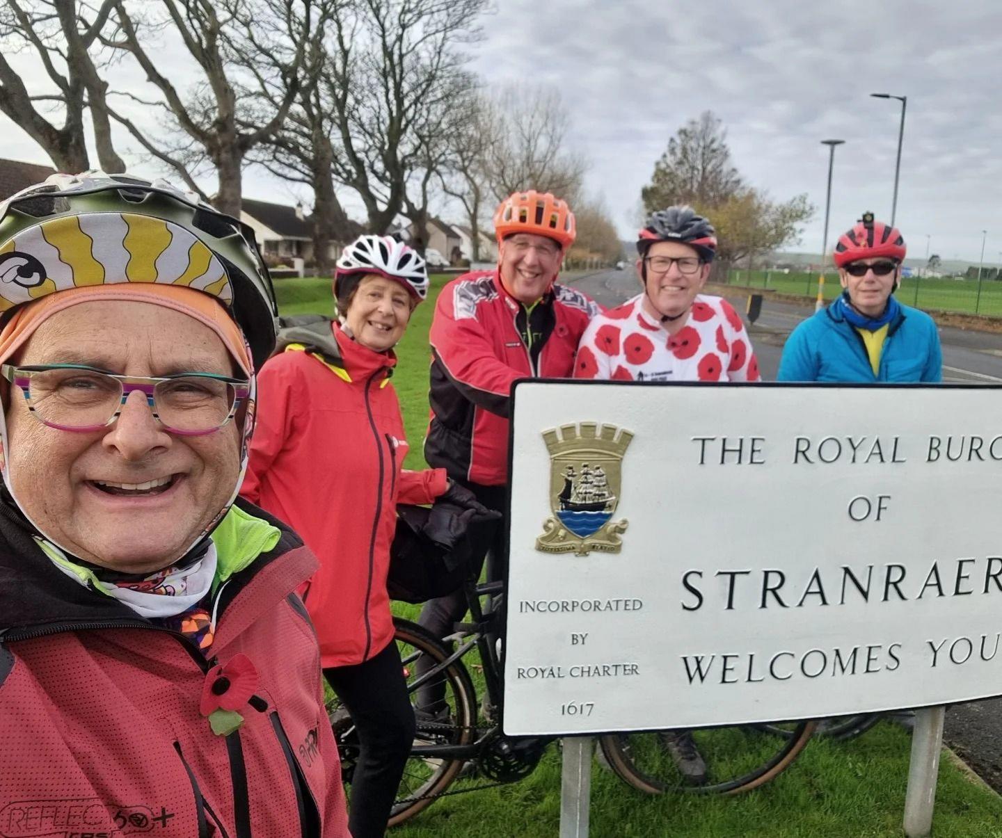 Timmy Mallett in cycling gear in the foreground stands in front of a sign welcoming people to Stranraer. Behind him are four people also in brightly coloured cycling attire standing behind the sign.