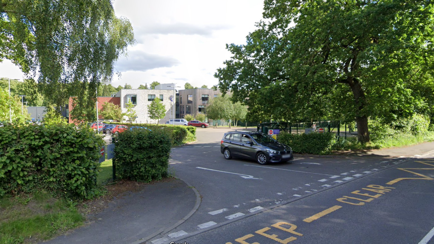 A driveway off a road leads towards a number of modern buildings in the distance. The road is lined with trees and hedges.