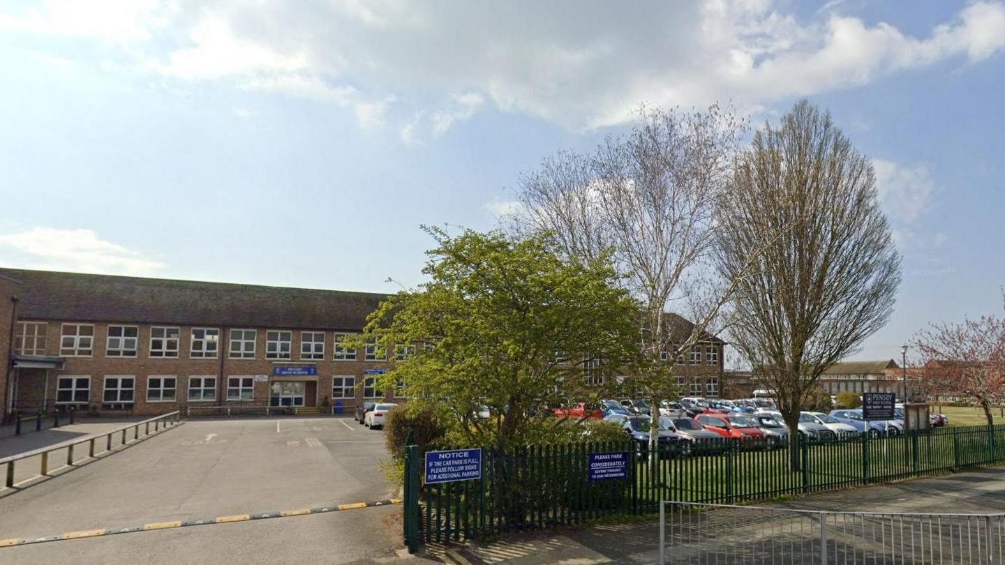 Front gate of Pensby High School with tree in the foreground and metal fences running along a path. 