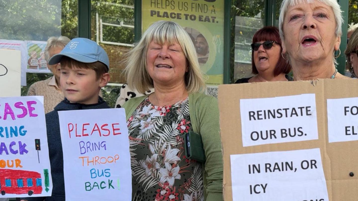 A group of protestors, holding signs calling for bus services to return to the village
