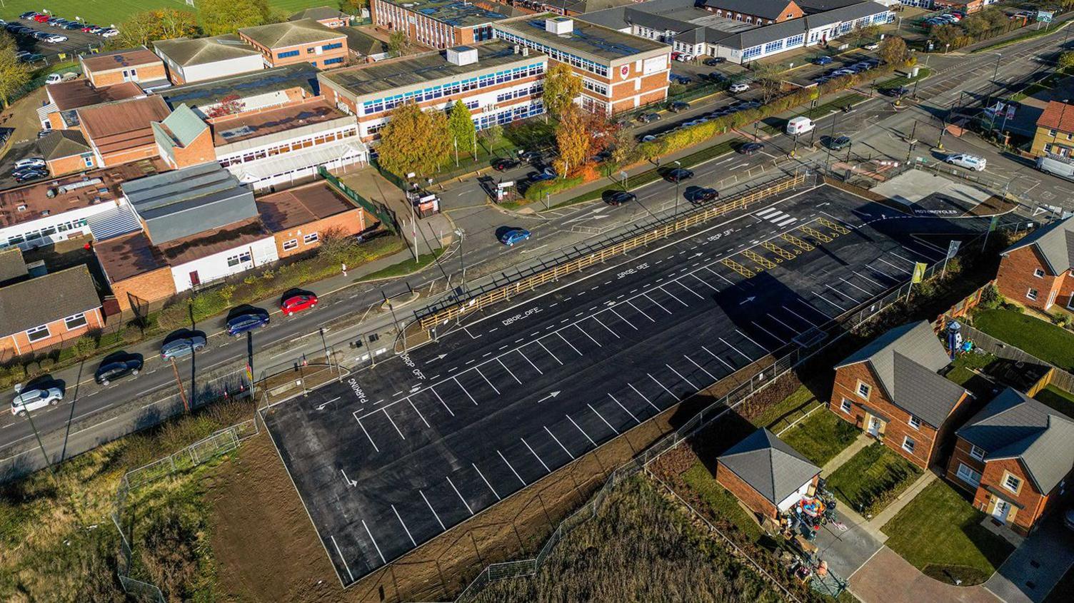 An aerial view of a rectangular black tarmac car park with white-marked parking bays. It is in the middle of an urban area with a school opposite and two rows of houses behind it.