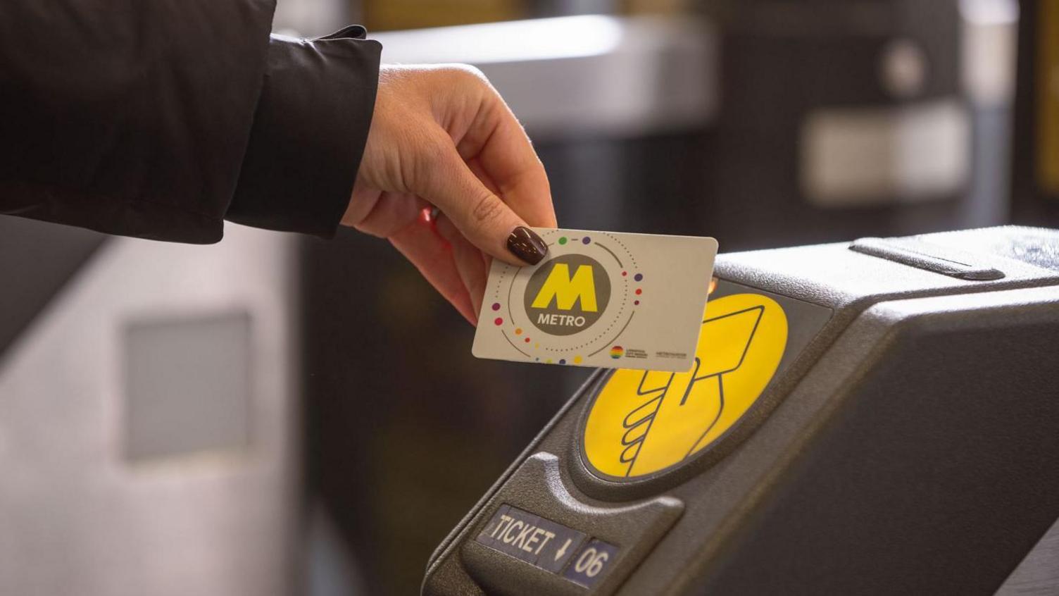 A closeup of a woman's hand holding a plastic card against a ticket barrier. The woman is wearing a black jacket and dark purple nail varnish. The card is white with a grey circle and a large, yellow letter M and white writing which reads METRO.