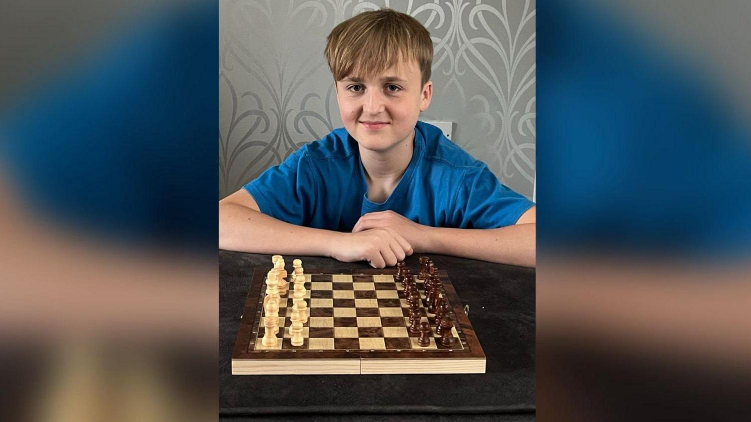 Calum (a boy with long brown hair and and a blue t-shirt) looks directly into the camera with sitting behind a chessboard