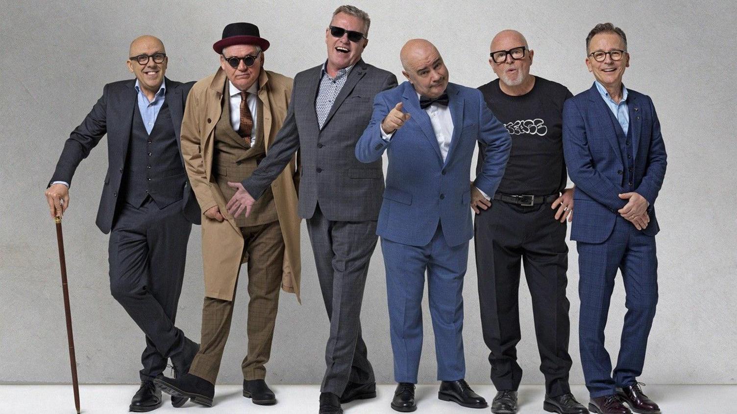 Six men are stood in a line against a plain grey background, dressed in formal  clothing. From left to right: a man in a dark suit with a light blue shirt and glasses holding a cane, another man in a tan trench coat over a suit with a hat and sunglasses, a man in a grey checkered suit wearing sunglasses, a man in a blue suit with a bow tie pointing a finger, a man in black trousers and black t-shirt with glasses and hands on hips, and another in a blue suit with glasses and hands resting on their front.