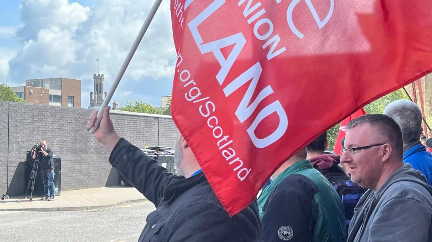 People protesting outside a Stagecoach depot with a Unite union red banner. There is a photographer taking pictures across the street