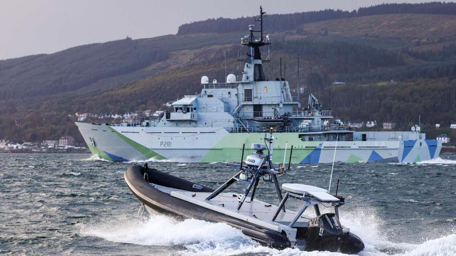 A Rattler boat sails through the water. It looks like a small inflatable boat, with a black inflatable rim around the edge and white interior. In the distance is a larger white naval vessel, which has some light green and blue angular patterns painted on the sides. In the distance of the sailing vessels is dark green mountainside.