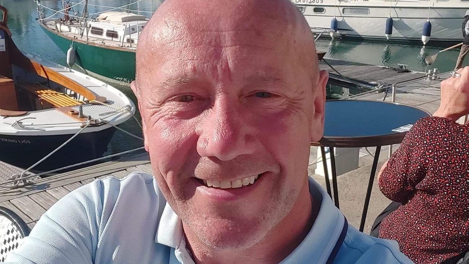 Brian Darby smiles at the camera. He is bald, has a tanned face and is sitting next to a marina.