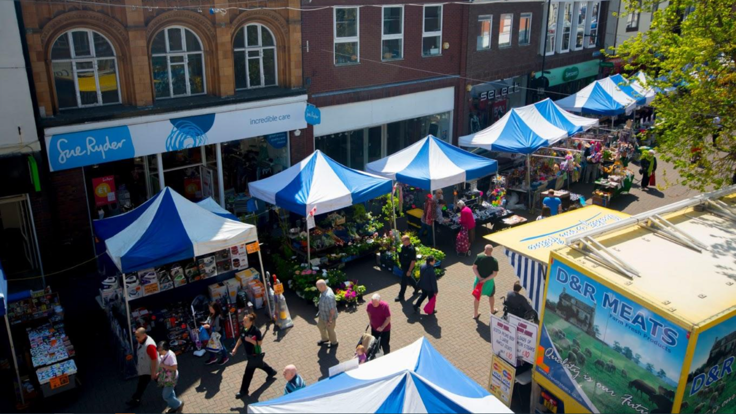 A market with blue and white stall lining a street, bathed in sunshine, with a parade of shops behind, including Sue Ryder, and opposite a butcher's stall, with customers walking past.