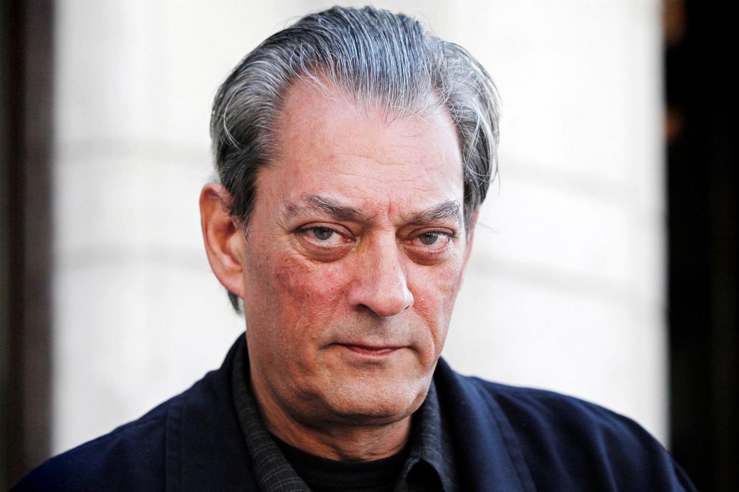 Paul Auster stares intently and seriously at the camera in this shot. He has slicked back grey hair. He appears to be outdoors and is wearing a dark jacket and dark shirt over a black T-shirt