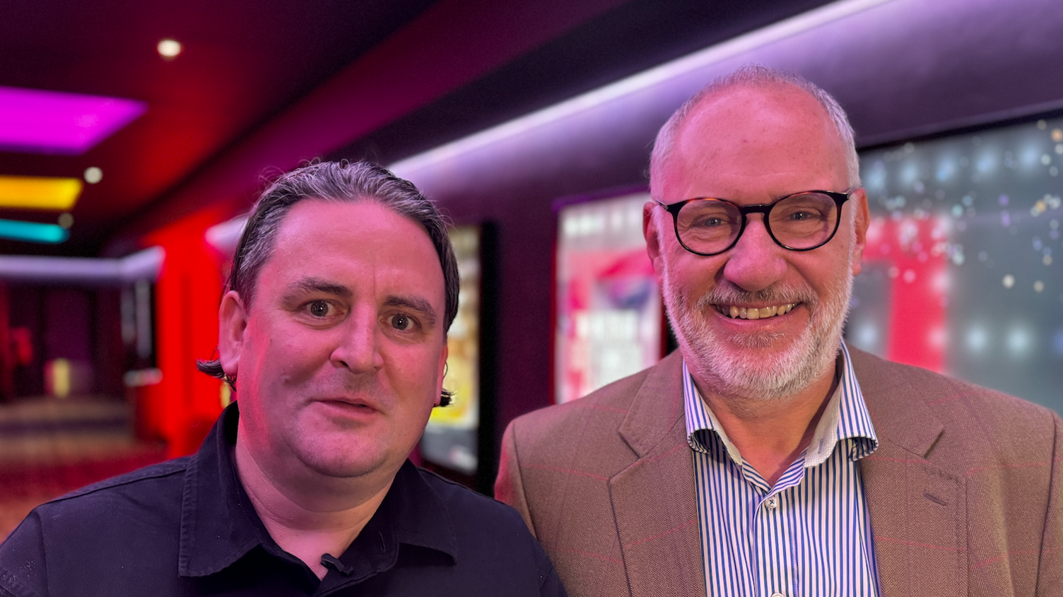 A man wearing a black shirt stands next to a man wearing a blazer with black rimmed glasses