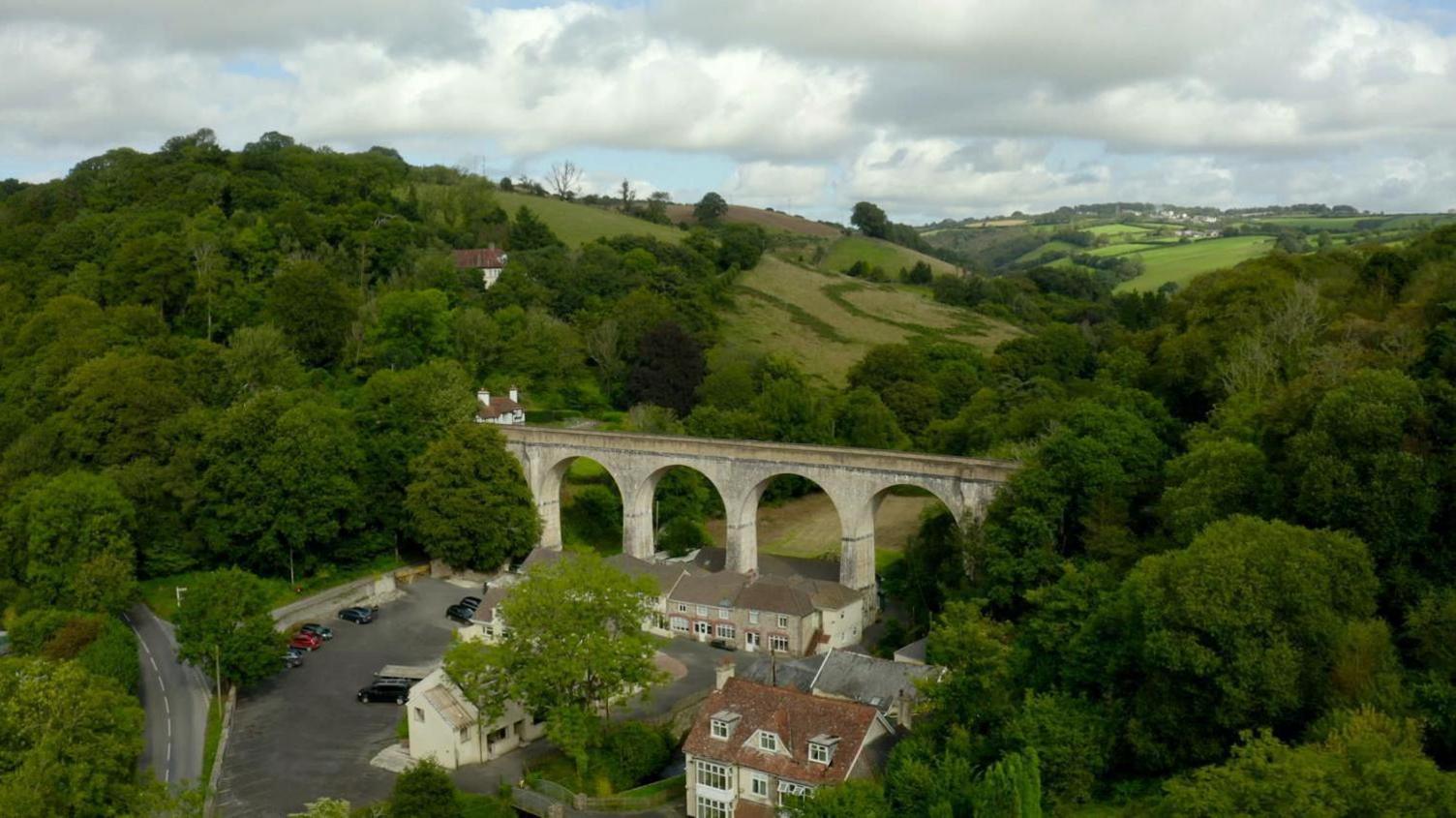 A drone photo of the mill in the countryside. A very scenic background with lots of trees and fields in the foreground. There is a viaduct in the middle of the picture with houses and a car park below. 