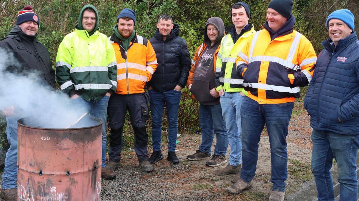 A group of men stand smiling around a red barrel with smoke coming out of it