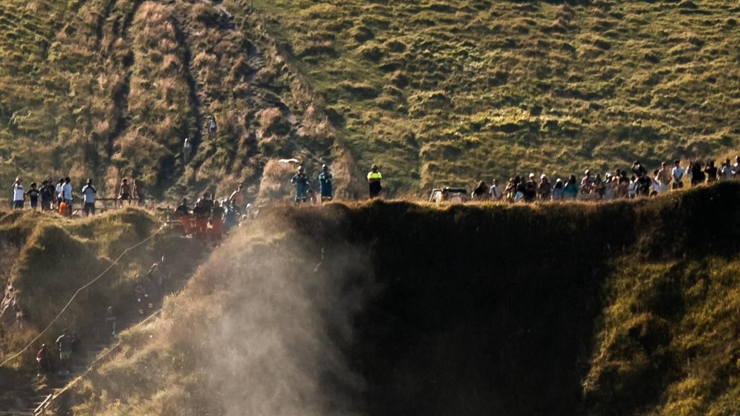 A line of people including some emergency crews lining the top of a cliff