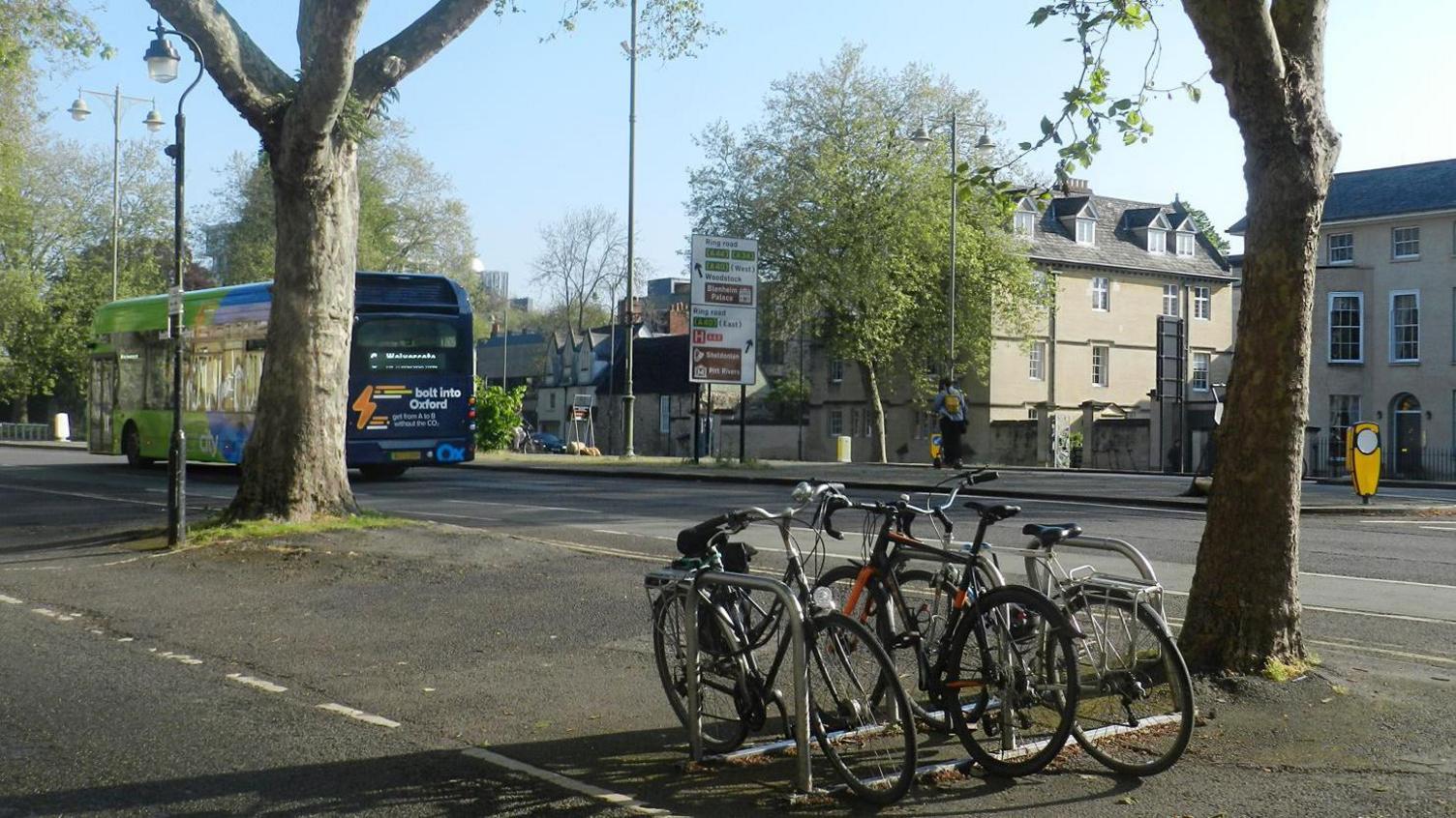 A cycle stand with three bikes parked under two trees on St Giles' boulevard in Oxford. A bus is heading north. It's a sunny day.